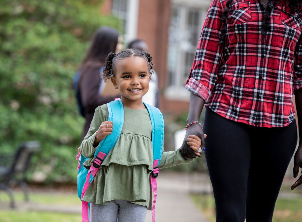 Girl wearing backpack walks into child care center holding hands with a parent