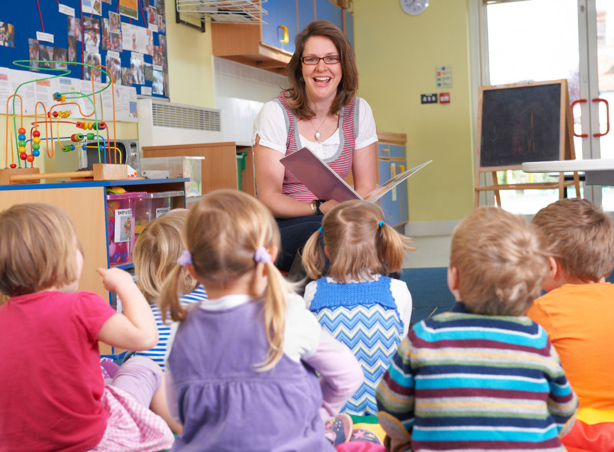 Young children sitting on the floor in a classroom listening to a teacher read