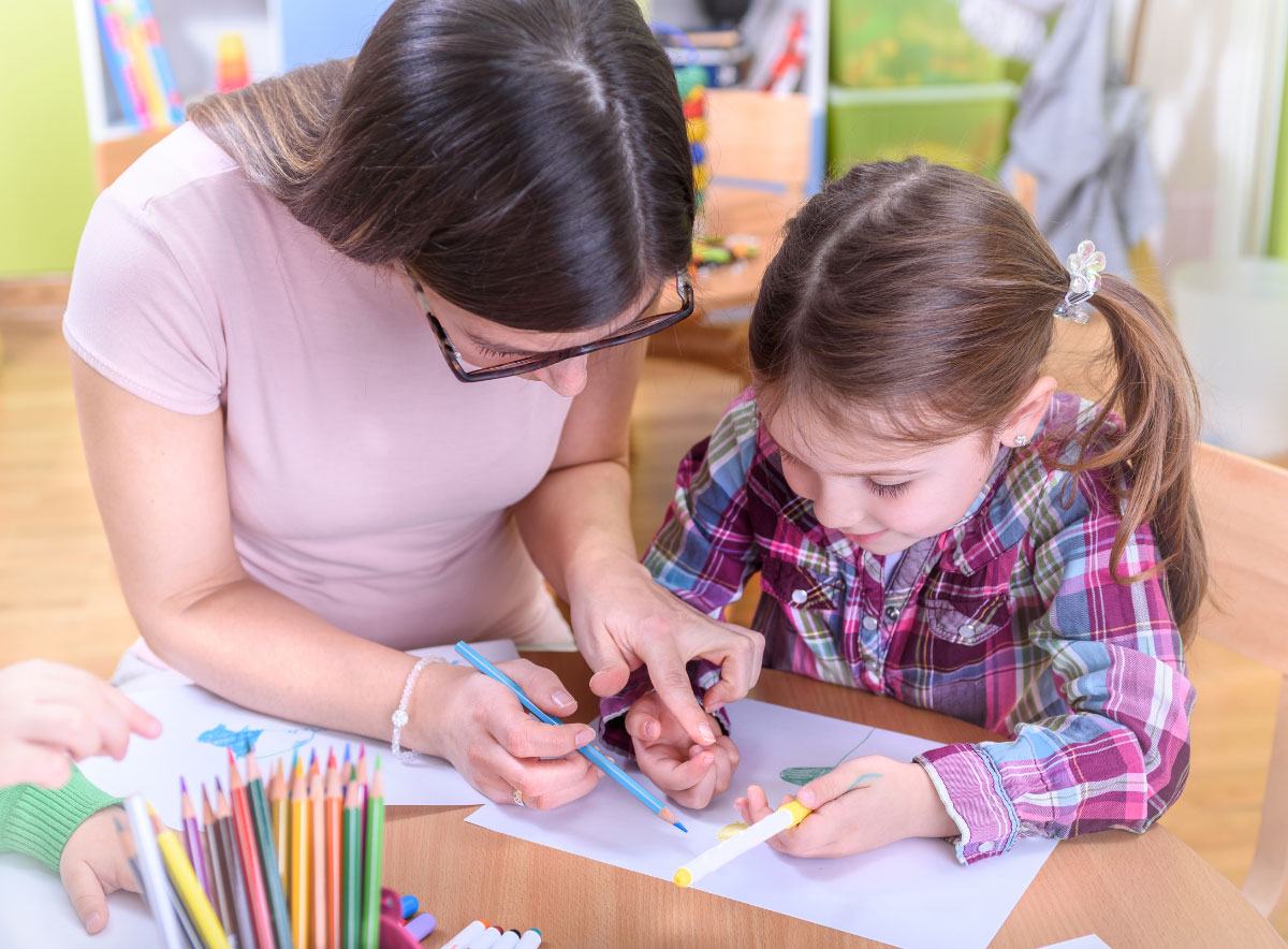 Teacher sitting with young girl student helping with color pencils at a child care center