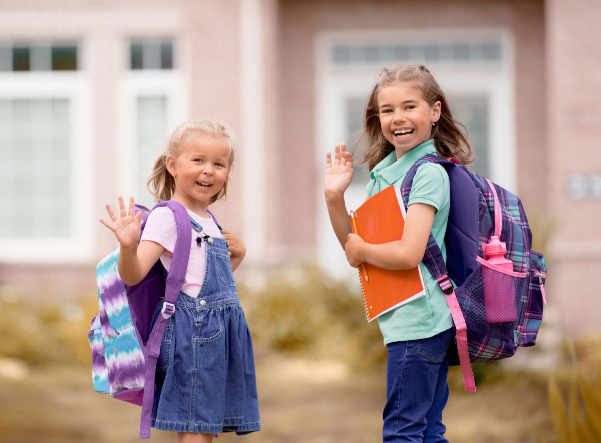 Two school age girls wearing backpacks and waving going into school at the end of summer