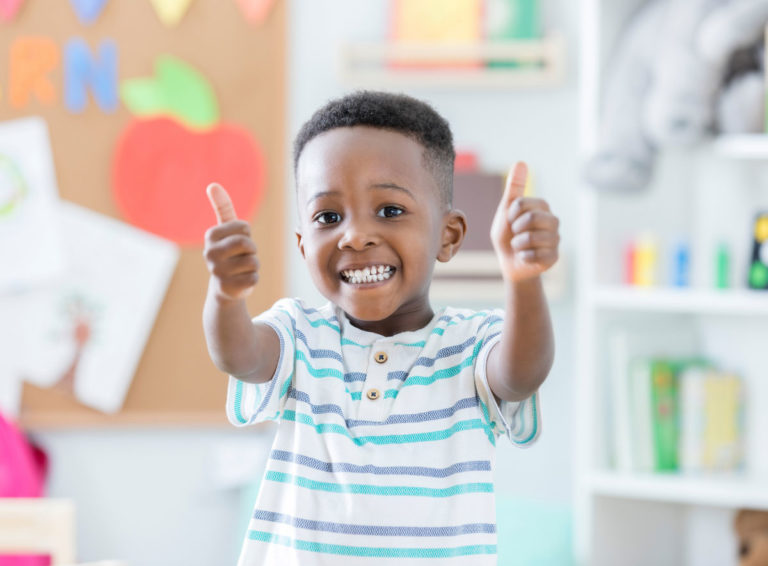 Young boy stands smiling with both thumbs up while in a child care center
