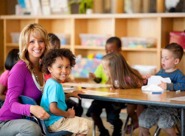 Smiling child care teach squats next to seated happy child at a table with students