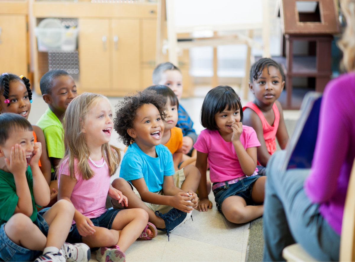 Children sit on the floor laughing excitedly while looking at a teacher with a book