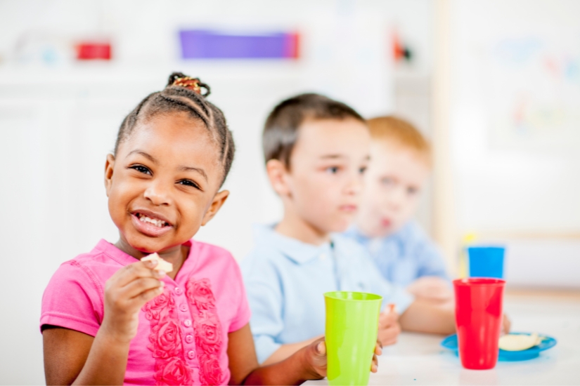 A girl smiles at the camera during a snack time as her peers sit nearby.