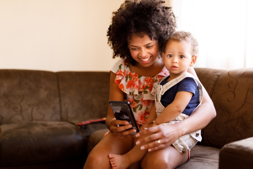 A mother holds her baby while smiling and looking at her phone.