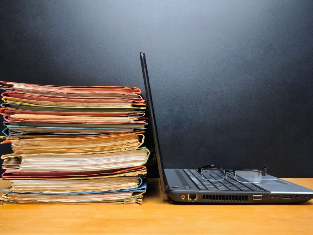 An open laptop sits next to a stack of paper files.