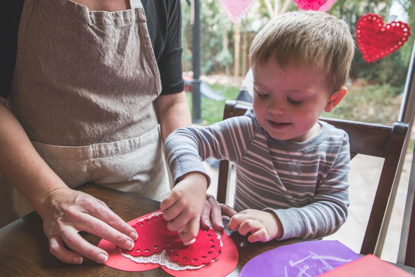 A young boy crafts a heart shaped card with help from a teacher.